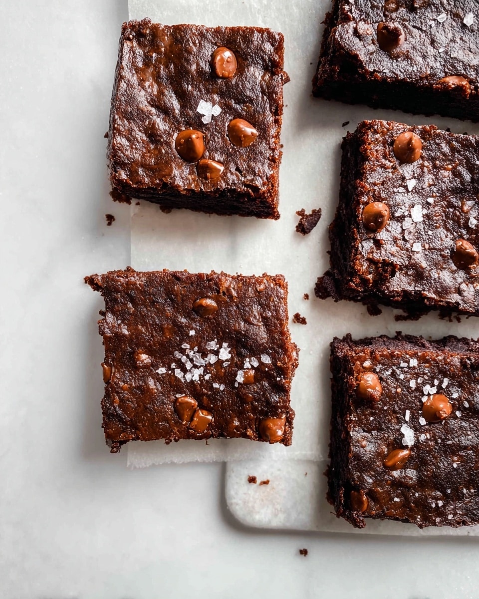 Four square brownie pieces sit on a white marbled surface with white parchment paper underneath. Each brownie has a dark brown top layer with a slightly shiny and cracked texture, dotted with melted chocolate chips and small flakes of sea salt. The rich, dense middle layer is visible on the edges, showing a moist and fudgy texture, darker in color than the top. Some crumbs are scattered around the brownies on the white marbled surface. photo taken with an iphone --ar 4:5 --v 7