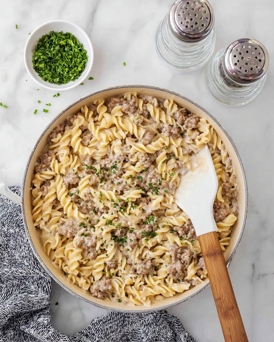 The image shows a white pan filled with creamy pasta mixed with browned ground meat. The pasta is light beige and twisted, evenly coated in a thick, creamy sauce, with small bits of green herbs sprinkled on top. A white spatula with a wooden handle is resting inside the pan, partly under the pasta. In the background, there is a small white bowl of chopped green herbs and two clear glass shakers with metal tops placed on a white marbled surface. A patterned gray and white cloth is at the bottom left corner. Photo taken with an iphone --ar 4:5 --v 7
