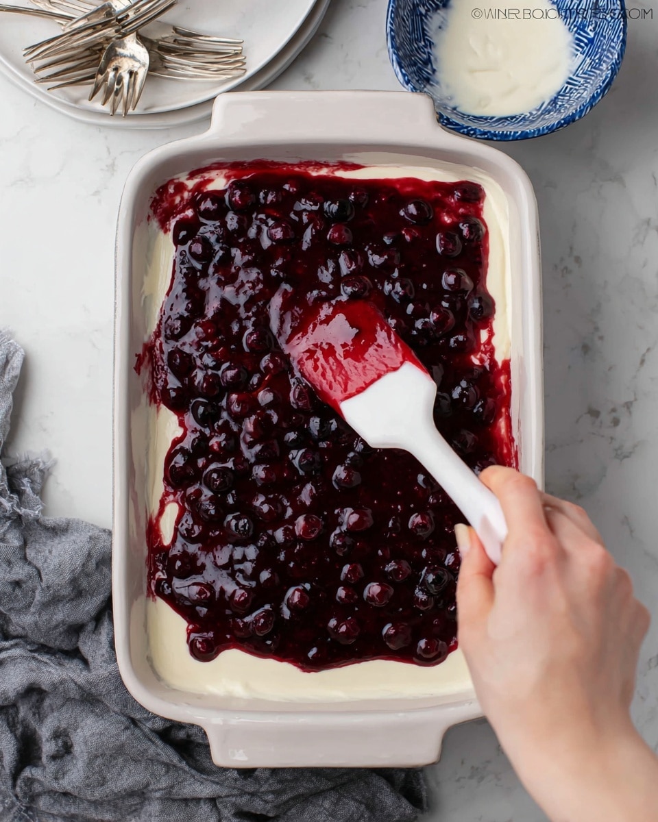 A rectangular white dish contains two visible layers: the bottom layer is smooth and creamy white, while the top layer is a thick, glossy dark red berry sauce with small whole berries scattered throughout. A woman's hand holds a white spatula with red berry sauce on it, spreading the sauce evenly over the creamy base. The dish is set on a white marbled surface, with a gray cloth partially visible on the lower left side and a blue and white bowl with more cream or sauce placed on the upper right. Nearby, stacked white plates and silver forks rest on the top left corner. photo taken with an iphone --ar 4:5 --v 7