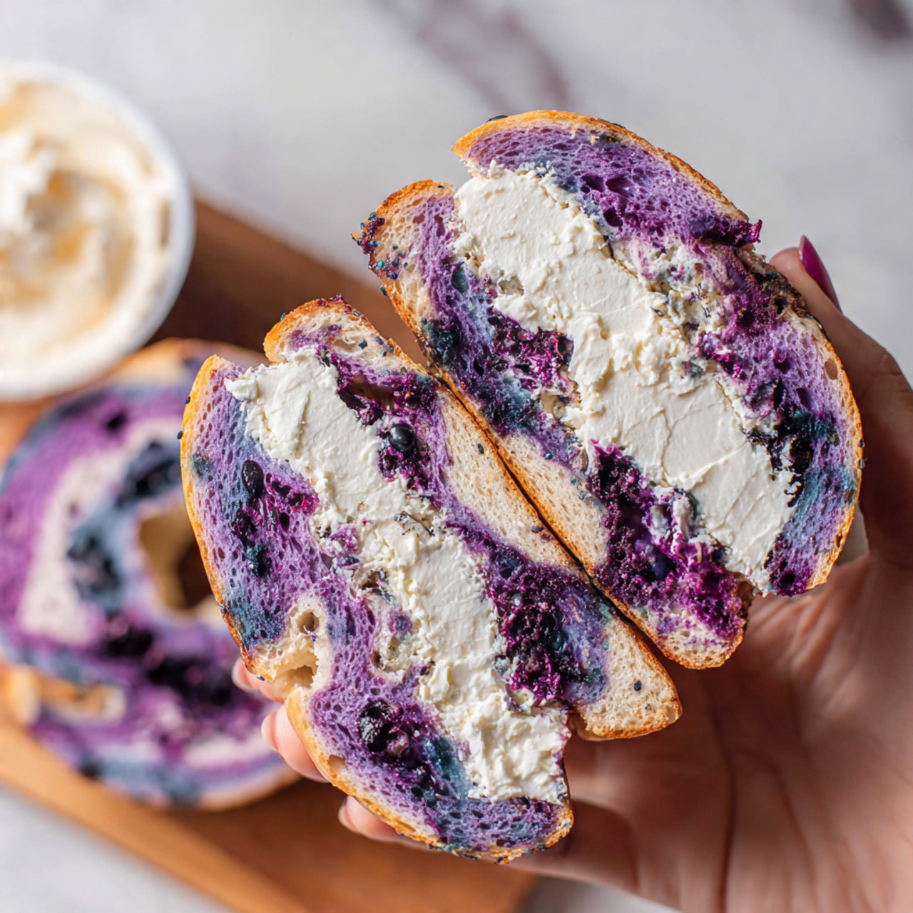 A close-up image of a sandwich held by a woman's hand, showing two halves of a purple swirl bagel with visible blueberry swirls throughout the soft, chewy bread. The bagel is sliced horizontally and packed with a thick, generous layer of white cream cheese spread in the middle. The background includes a white marbled surface with a wooden board, and the top half of the bagel is set down next to the sandwich along with an open container of cream cheese. The photo taken with an iphone --ar 4:5 --v 7
