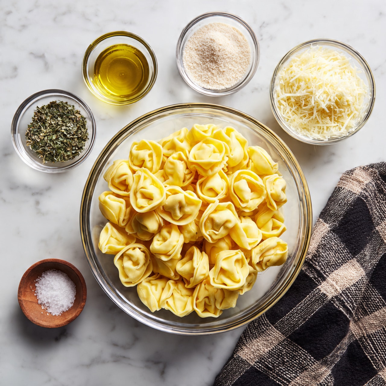 A clear glass bowl filled with yellow tortellini pasta sits in the center on a white marbled surface, surrounded by four small clear glass bowls containing various ingredients: one with green dried herbs at top left, one with olive oil at top right, one with pale beige powder at middle left, and one with grated cheese at middle right, plus a small bowl of salt near the bottom left. A black and tan checkered cloth is loosely placed to the right near the bowl of cheese. photo taken with an iphone --ar 4:5 --v 7
