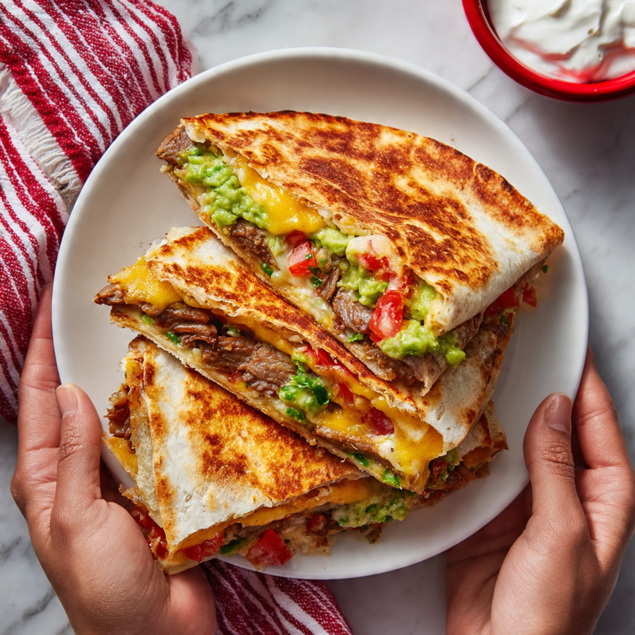 A pair of hands hold a quesadilla cut in half over a white plate, showing its four visible layers from top to bottom: a toasted, golden-brown tortilla, a layer of melted yellow cheese, a layer of green guacamole with a bit of red tomato, and pieces of grilled brown meat at the bottom, with another soft white tortilla inside. The setting features a white marbled surface with a red and white striped cloth on the side and a red bowl of white sour cream nearby. Photo taken with an iphone --ar 4:5 --v 7