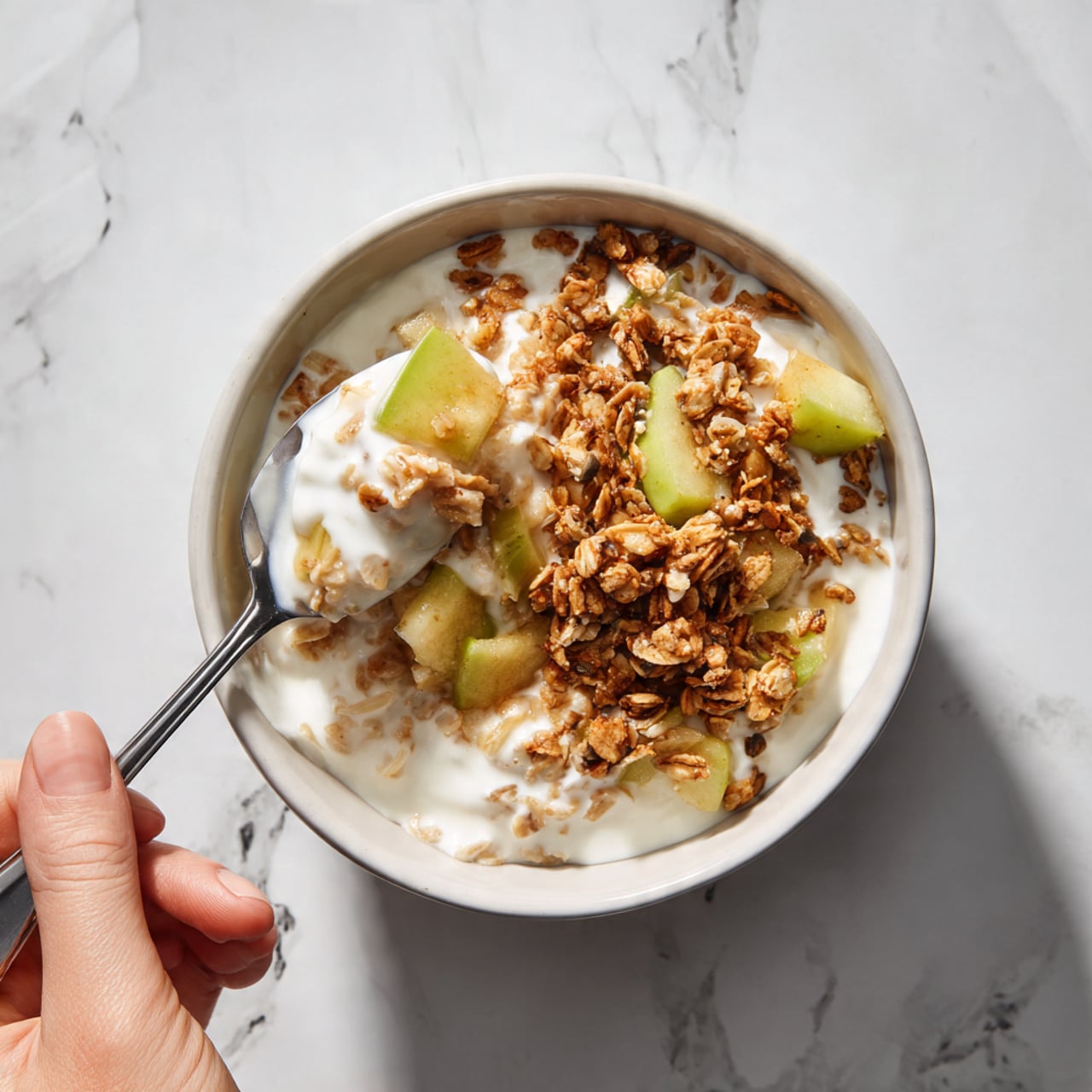 A white bowl filled with three main layers: the bottom layer looks like cooked oats mixed with pieces of soft, light green apple, the middle layer has crunchy golden and toasted brown granola clusters scattered throughout, and the top layer is a glossy, thick white sauce or yogurt draping over the oats and granola, giving a creamy texture. A spoon lifts a spoonful of the mix, showing the oats, apple pieces, and granola together, with steam faintly rising. A woman's hand is holding the spoon, and the background is a white marbled texture. photo taken with an iphone --ar 4:5 --v 7