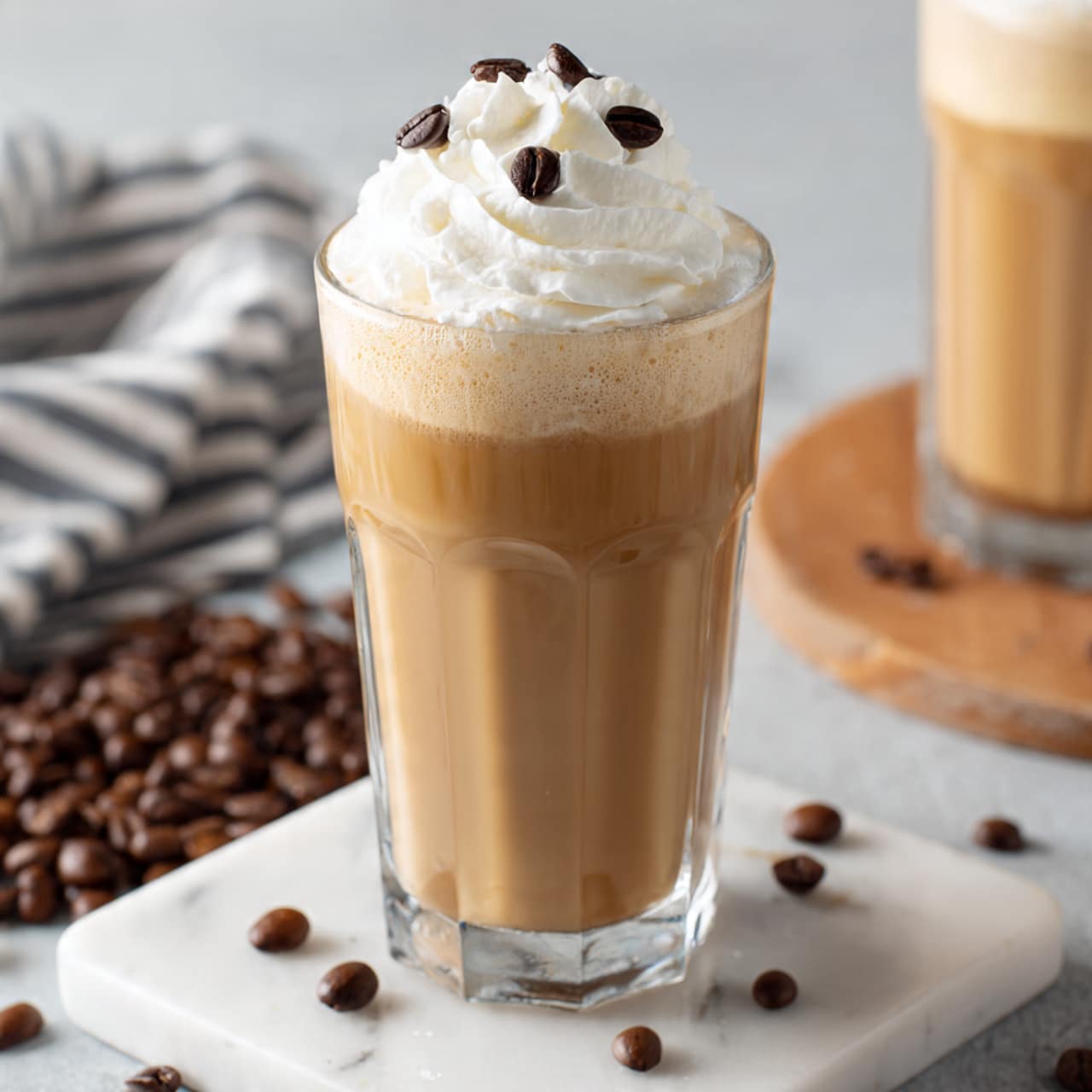 A tall clear glass filled with a creamy light brown coffee drink showing a smooth, slightly frothy texture. On top, there is a thick dollop of white whipped cream, which is decorated with several small dark brown coffee beans. The glass sits on a white marbled surface with scattered coffee beans around it, with a blurred background showing part of another similar glass and a wooden board. A striped cloth lies nearby, adding a cozy touch. photo taken with an iphone --ar 4:5 --v 7