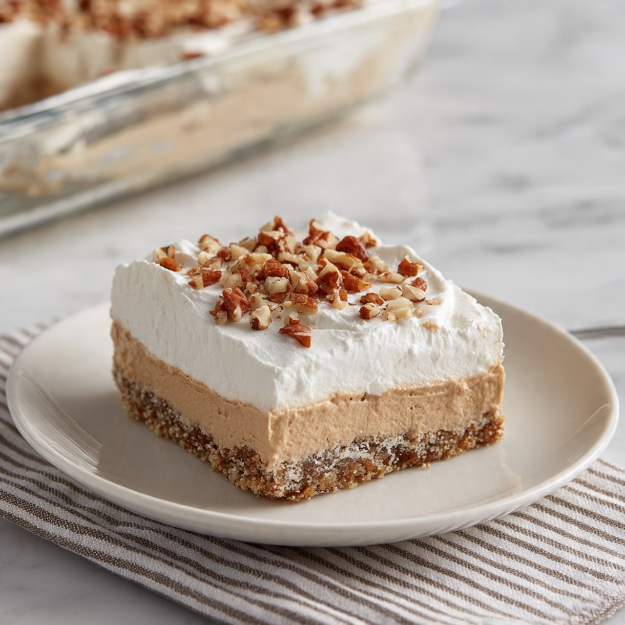 A square piece of dessert is placed in the center of a white plate. The dessert has three clear layers: the bottom layer is a light, crumbly crust with a rough texture; the middle layer is smooth and light brown, creamy in look; the top layer is thick white whipped cream spread evenly with soft curves. Small chopped nut pieces are sprinkled over the whipped cream, adding texture and color contrast. In the background, part of a glass baking dish with the remaining dessert is visible on a white marbled surface with a striped cloth underneath. Photo taken with an iphone --ar 4:5 --v 7