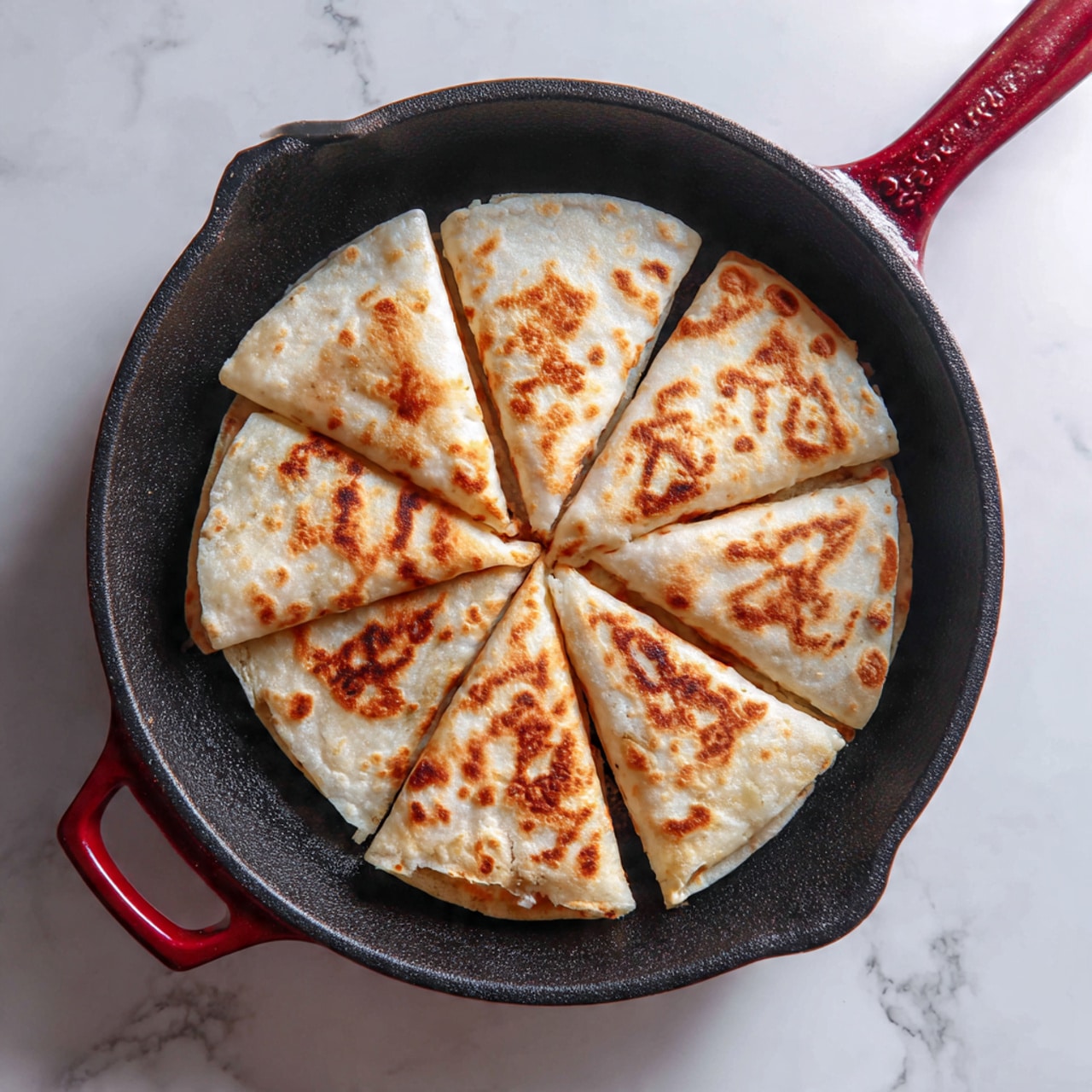 A folded tortilla is placed in the center of a black cast iron pan with a red handle. The tortilla has six overlapping flaps folded over the filling, creating a star-like pattern with a round, toasted brown spot in the middle. The rest of the tortilla is light beige with some toasted light brown areas, showing a soft and slightly uneven texture. The pan sits on a white marbled surface. photo taken with an iphone --ar 4:5 --v 7