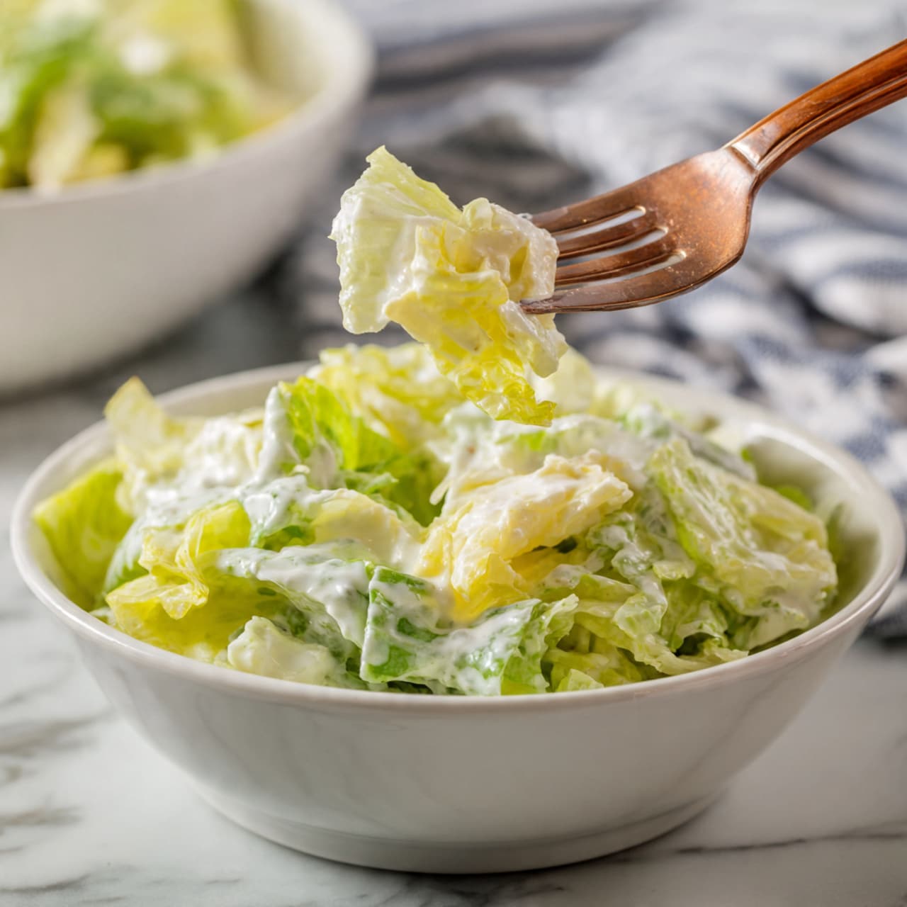 A close-up image of a white bowl filled with creamy salad, showing several layers of pale green and light yellow lettuce coated in a white, creamy dressing; a fork is lifting a small portion of the salad above the bowl, highlighting the mix of soft, wilted leaves and fresh, crisp pieces, set on a white marbled surface with a blurred background featuring a white bowl of more salad and a striped cloth. photo taken with an iphone --ar 4:5 --v 7