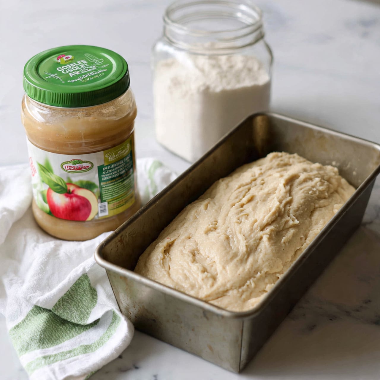 A metal loaf pan filled with a thick, pale beige batter showing a slightly rough and wet texture on top, placed on a white marbled surface. Next to the loaf pan is a nearly full clear plastic jar of chunky apple sauce with a green lid and green label featuring a bright red apple image. Behind these items, there is a clear container filled with white sugar. A white cloth with green stripes lies partially under the apple sauce jar. The setting looks like a baking preparation area. Photo taken with an iphone --ar 4:5 --v 7