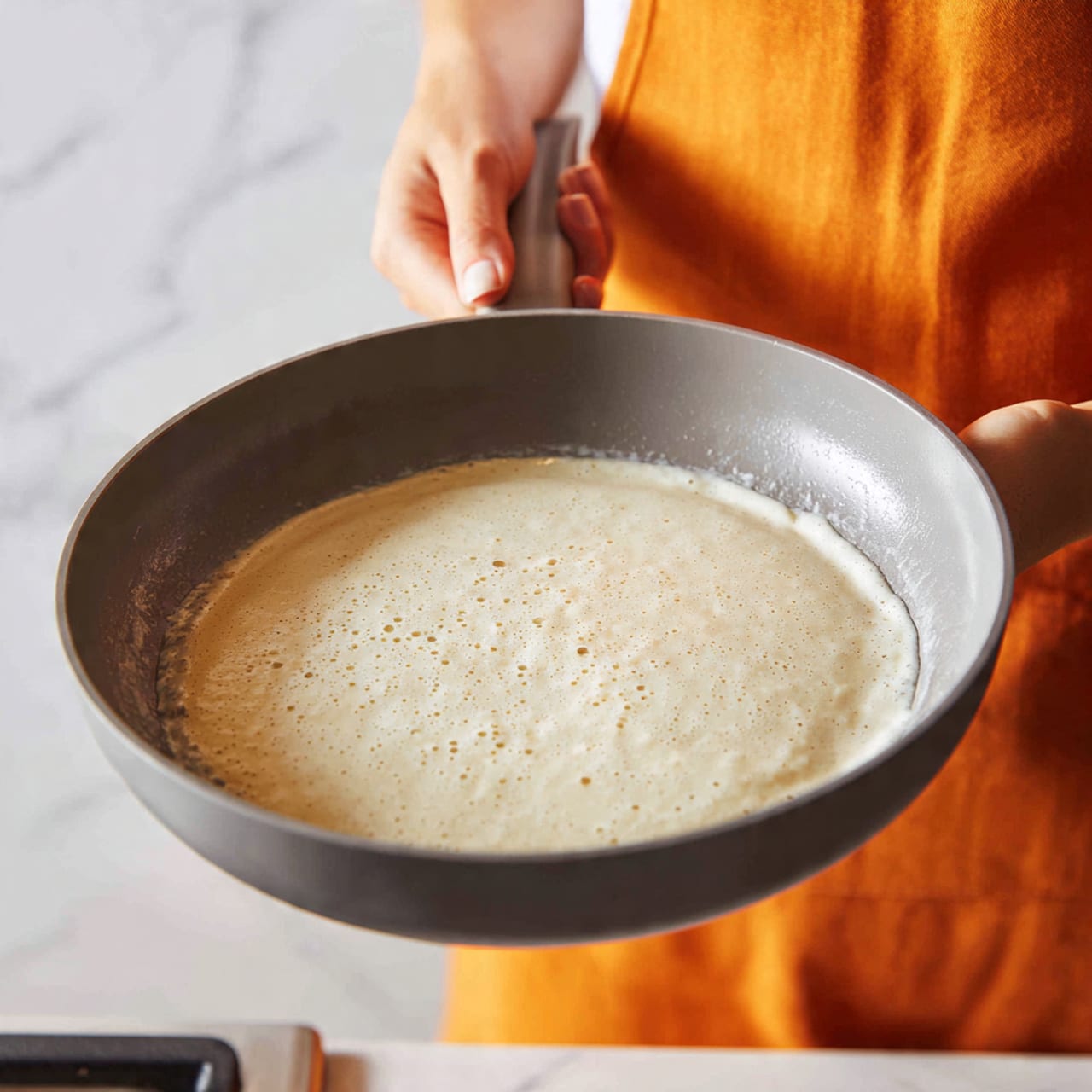 A close-up image of a gray non-stick frying pan being held with a woman's hand over a stove. Inside the pan, there is a single light cream-colored batter layer almost covering the bottom, with some uneven textures and tiny bubbles forming on the surface. The background is a white marbled countertop, and the person is wearing an orange apron. Photo taken with an iphone --ar 4:5 --v 7