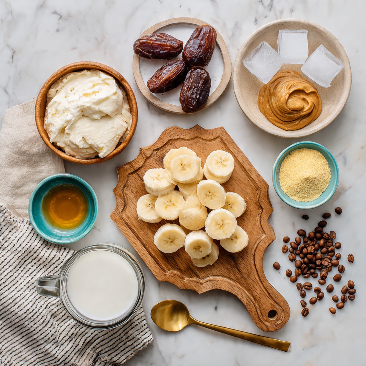 The image shows several ingredients arranged neatly on a white marbled surface and a wooden cutting board with scalloped edges. There is a small glass bowl filled with frozen banana slices in the center of the cutting board. To the left, a wooden bowl holds a creamy white substance, and below it, a tiny glass bowl contains a dark liquid. To the right of the frozen bananas on the cutting board, a small white bowl contains golden honey. Next to it is a turquoise bowl filled with a pale yellow powder. Above the cutting board on the white marbled surface are two dark dates resting on a small white plate and a beige scalloped bowl with thick light brown almond butter. To the right, there is a white bowl full of ice cubes on top of a black and white striped cloth. In the lower right corner, a clear glass pitcher holds milk, and coffee beans are scattered around it. A golden spoon lies horizontally in front of the cutting board. photo taken with an iphone --ar 4:5 --v 7