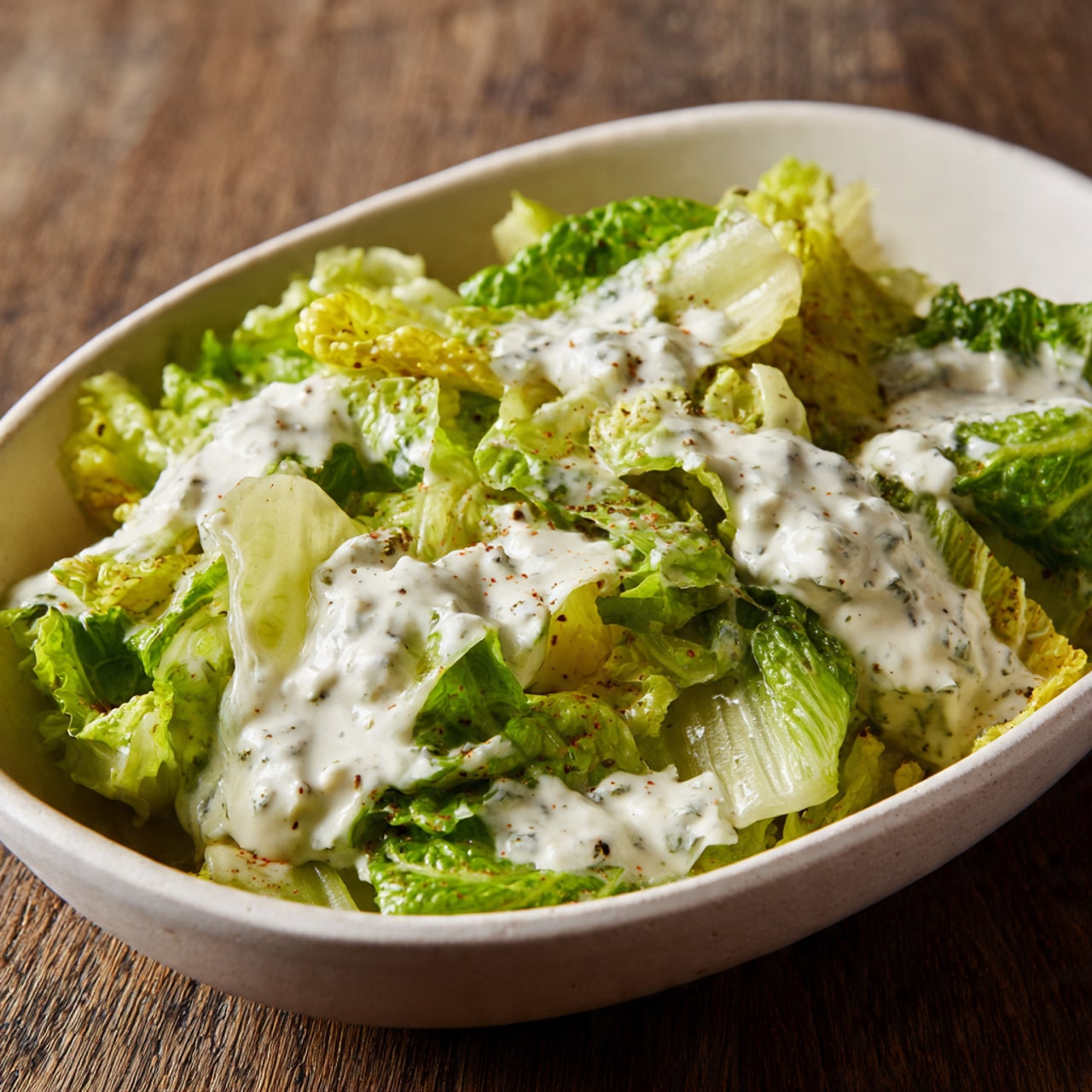 A white oval bowl filled with a base layer of chopped light green and pale yellow lettuce leaves, slightly crisp and layered evenly throughout. On top, there are uneven dollops of creamy white dressing with visible small chunks and specks, spread in a scattered pattern mostly along the center and upper right area of the lettuce. The bowl is placed on a brown wood-textured surface. The texture of the dressing looks thick and slightly lumpy over the fresh, leafy vegetables. Photo taken with an iphone --ar 4:5 --v 7