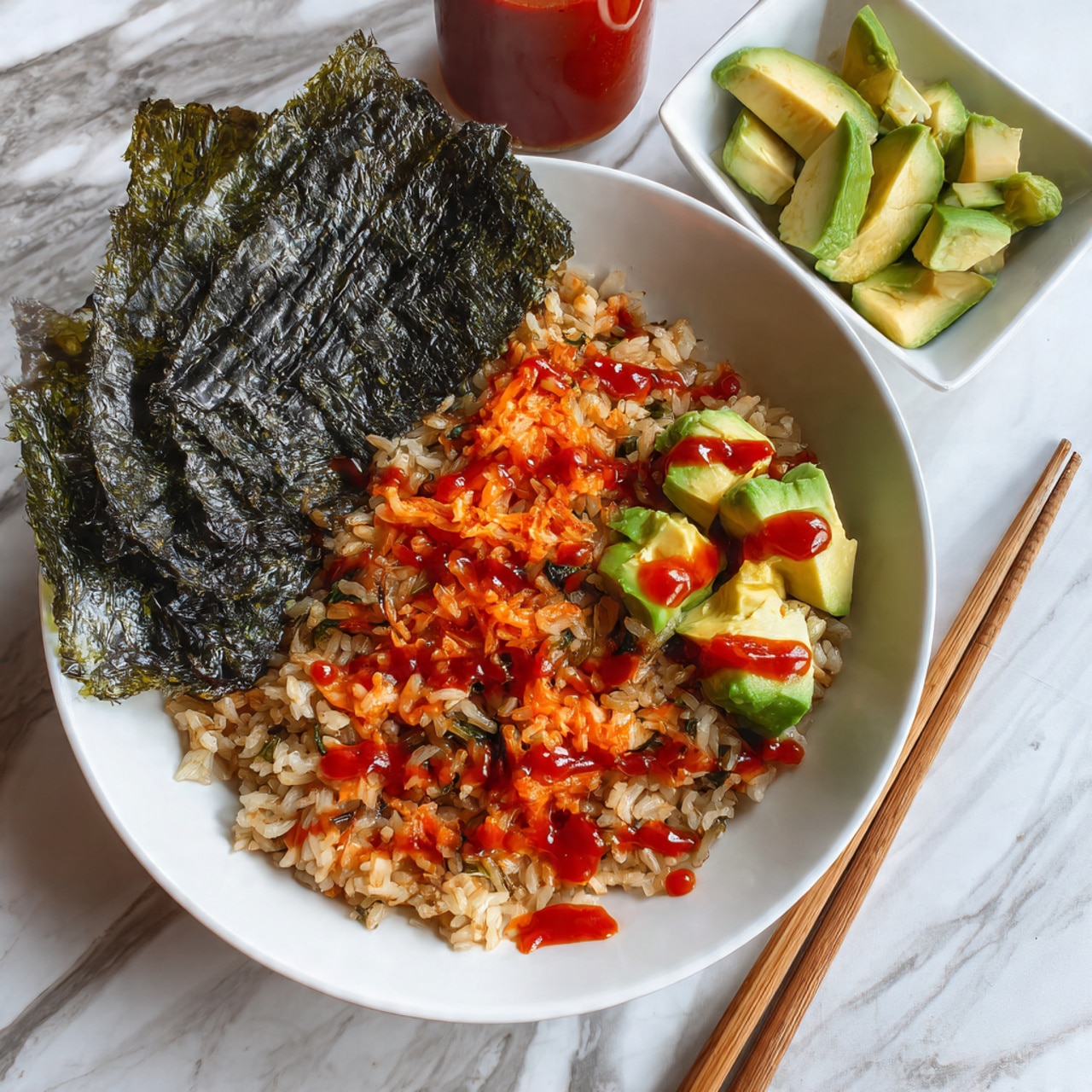 A white plate sits on a white marbled surface holding a dish with three clear layers: at the bottom, a layer of light brown cooked rice mixed with light-colored pieces of fish or tofu; on the right side, a pile of chopped light green avocado cubes; at the top left corner, a large dark green sheet of seaweed partly tucked under the rice. Bright red sauce is drizzled across the rice and avocado in small uneven drops, and some white sesame seeds are sprinkled over the rice. Two wooden chopsticks rest on the right edge of the plate. Photo taken with an iphone --ar 4:5 --v 7