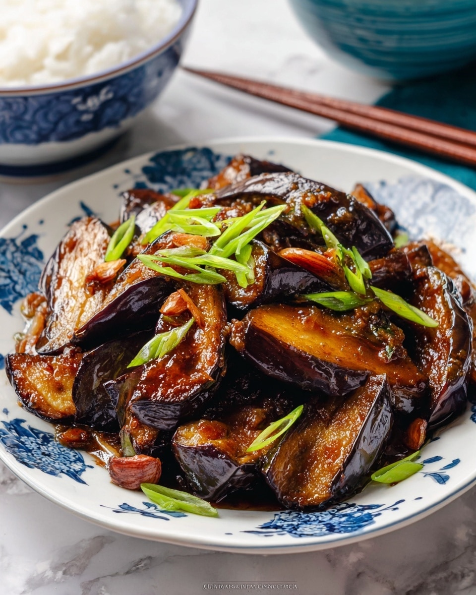 A white plate with blue flower patterns holds a pile of cooked eggplant slices, each with dark purple skin and soft, light brown inner flesh. The eggplants are covered in a shiny, dark brown sauce that makes some parts look glossy and rich. Small pieces of garlic are visible on the eggplants. Bright green sliced spring onions are sprinkled on top, adding a fresh contrast to the dish. The plate is placed on a white marbled surface with part of a blue and white bowl and wooden chopsticks seen blurry in the background. photo taken with an iphone --ar 4:5 --v 7