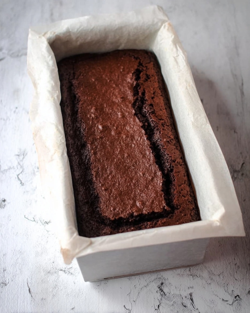 A rectangular brown chocolate cake sits in a white baking pan lined with white parchment paper that slightly folds over the sides. The cake has a slightly cracked and raised top with a textured surface showing darker and lighter brown tones, indicating moistness and richness. The pan rests on a white marbled surface, which adds a clean and bright contrast to the dark cake. photo taken with an iphone --ar 4:5 --v 7