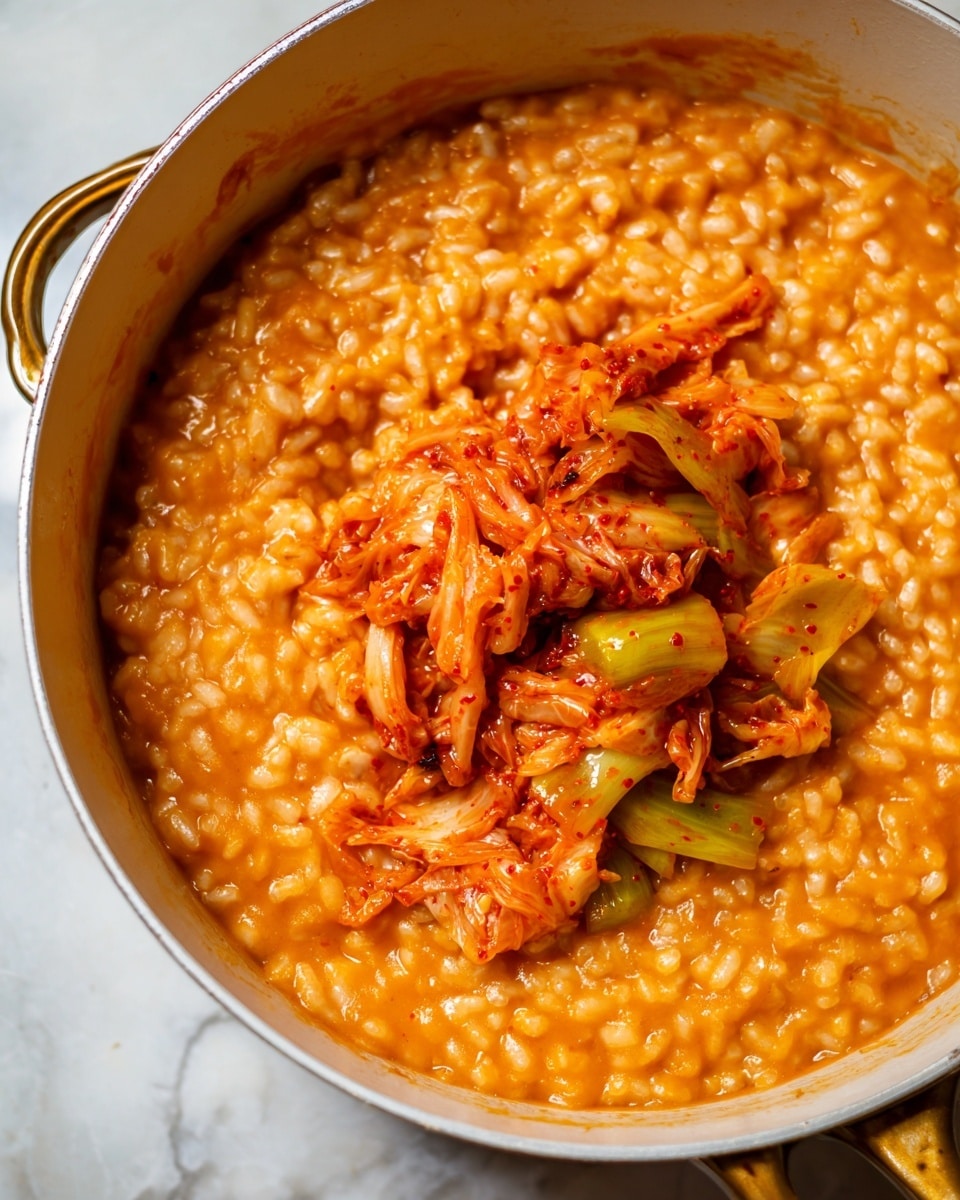 The image shows a white pot filled with thick, creamy orange risotto mixed with soft, small grains and sauce. On top, there is a layer of bright reddish-orange kimchi, with pieces of cabbage and shredded vegetables, adding texture and moisture. The pot has a golden handle and rests on a white marbled surface. The colors contrast well, with the orange risotto forming the base layer and the more vibrant kimchi sitting on top in the center. Photo taken with an iphone --ar 4:5 --v 7