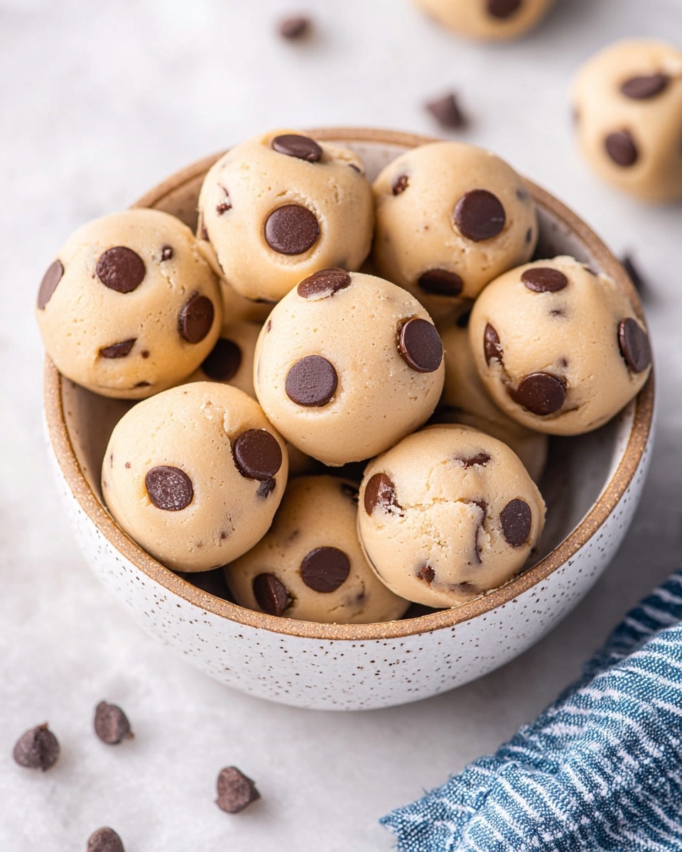 A close-up of a bowl filled with round, light beige cookie dough balls dotted with dark brown chocolate chips, each dough ball plump and smooth with three to four chocolate chips visible on their tops and sides, the bowl is white with a speckled texture and sits on a white marbled surface scattered with a few loose chocolate chips; a blue and white striped cloth is partially visible on the right side. photo taken with an iphone --ar 4:5 --v 7