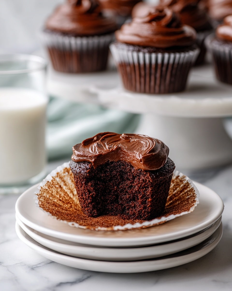 A small chocolate cupcake with a bite taken out of the front shows its soft, dark brown inside. It has a thick, shiny layer of dark chocolate frosting on top, spreading slightly over the edges. The cupcake sits on two stacked white plates placed on a white marbled surface. In the background, there are several whole chocolate cupcakes on a white plate and on a white cake stand, all with smooth, glossy dark chocolate frosting. A clear glass of milk is partially visible to the left. photo taken with an iphone --ar 4:5 --v 7