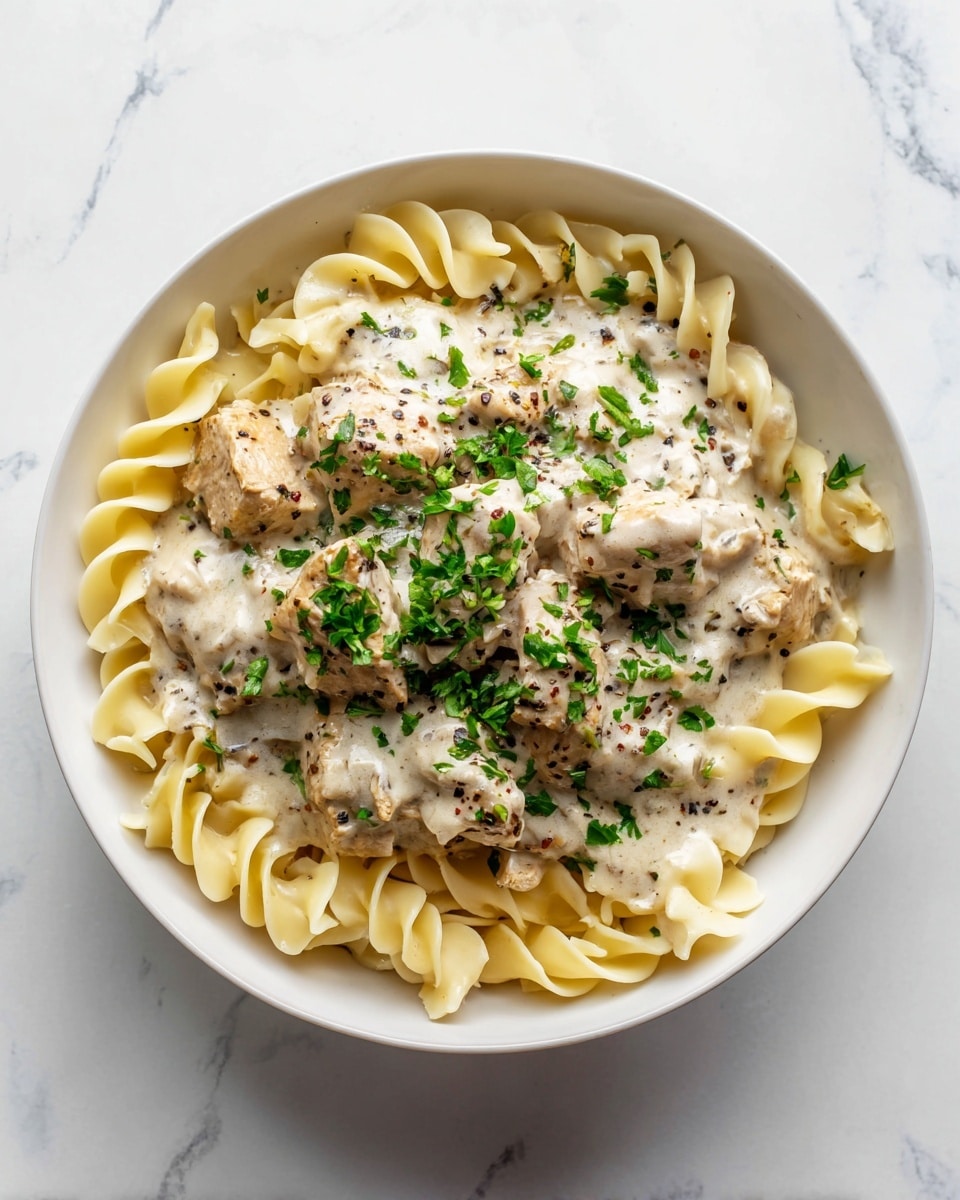 A white bowl filled with three layers: the bottom layer has pale yellow cooked pasta noodles with a smooth texture, loosely piled; the middle layer shows chunks of browned chicken cooked evenly, covered in a creamy light beige sauce speckled with black pepper; the top layer is sprinkled with small bright green chopped parsley leaves scattered over the sauce, adding a fresh contrast. The bowl sits on a white marbled surface. photo taken with an iphone --ar 4:5 --v 7