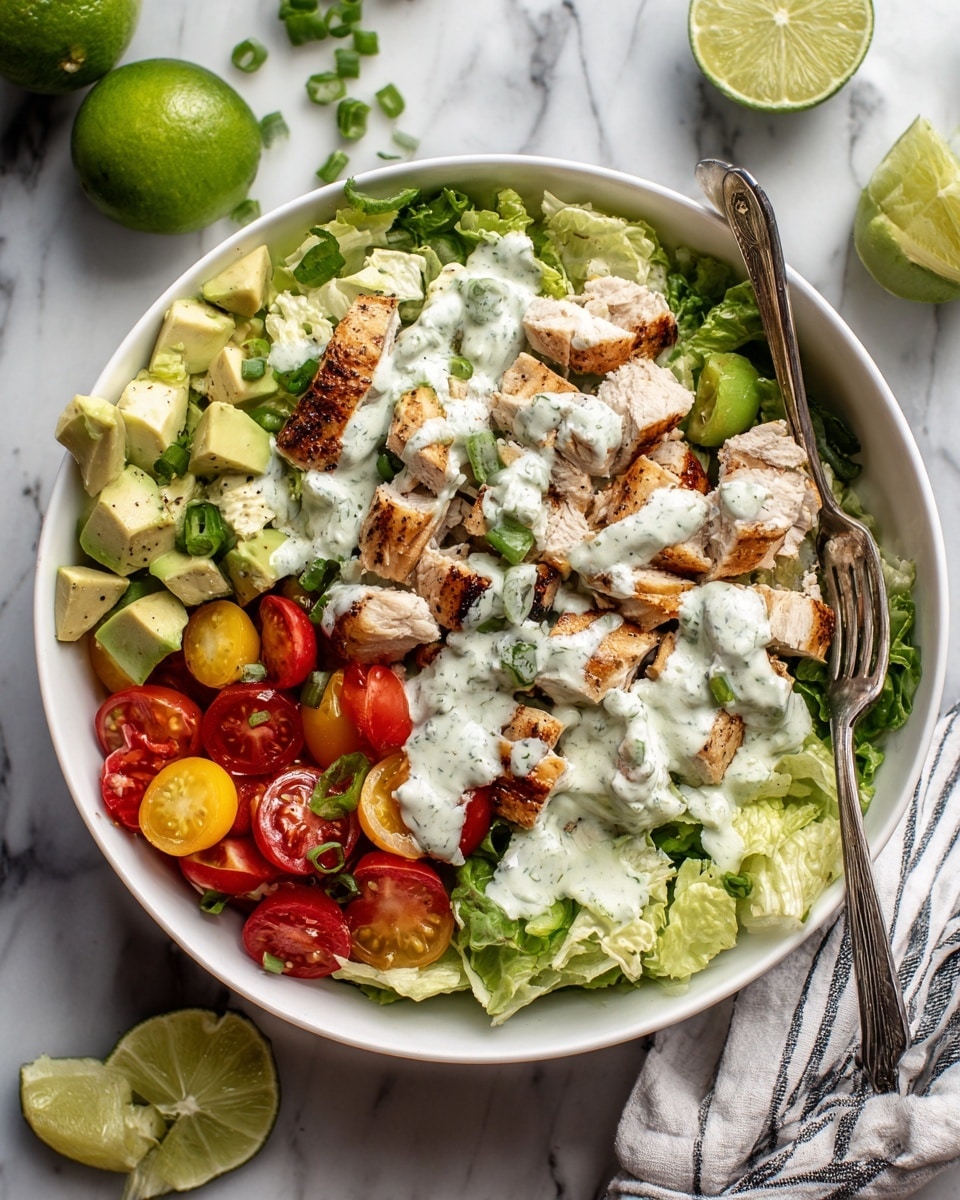 In a white bowl, there is a layered salad starting with a bed of fresh green lettuce at the bottom. On top of the lettuce, there are slices of bright red cherry tomatoes and light yellow avocado pieces spread around. Several chunks of light beige cooked chicken are arranged over these vegetables. The salad is topped with a creamy white sauce with green specks, drizzled generously over the chicken and vegetables. Bright green chopped scallions are sprinkled on top, adding extra color. There are two lime wedges placed on the side near the edge of the bowl, and a fork rests partially inside the bowl, touching the salad. The bowl sits on a white marbled surface with some limes and a cloth visible around it photo taken with an iphone --ar 4:5 --v 7