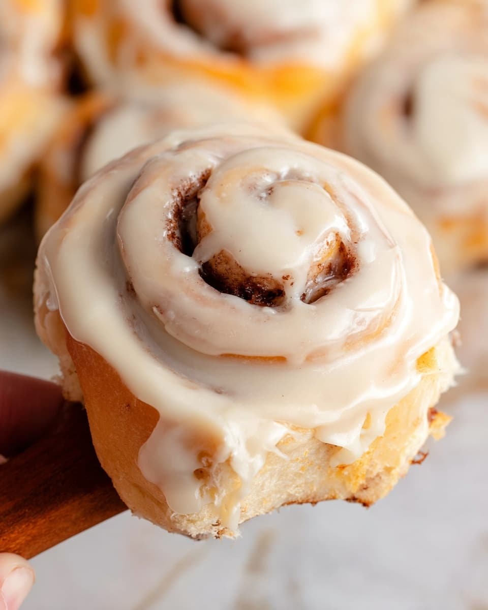 A close-up view shows a single cinnamon roll held by a woman's hand above a white marbled surface, with other cinnamon rolls visible blurred in the background. The roll has a visible spiral with one layer of light golden-brown dough mixed with a darker cinnamon filling. The whole top and sides are covered with a thick, smooth, creamy white icing that has a slightly glossy texture. The icing fills some crevices of the swirl and drips slightly over the edges, making the roll look soft and moist. photo taken with an iphone --ar 4:5 --v 7