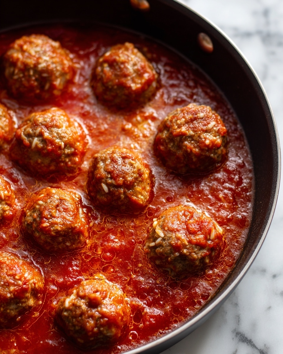 The image shows a close-up of round meatballs cooking in a rich, red tomato sauce. Each meatball has a rough texture with bits of white rice mixed in, and some are partially covered in the sauce. The sauce bubbles gently around the meatballs, creating small, glossy spots and a vibrant, thick liquid layer. The cooking vessel's edge is dark and contrasts with the bright red sauce. The background is a white marbled surface. Photo taken with an iphone --ar 4:5 --v 7