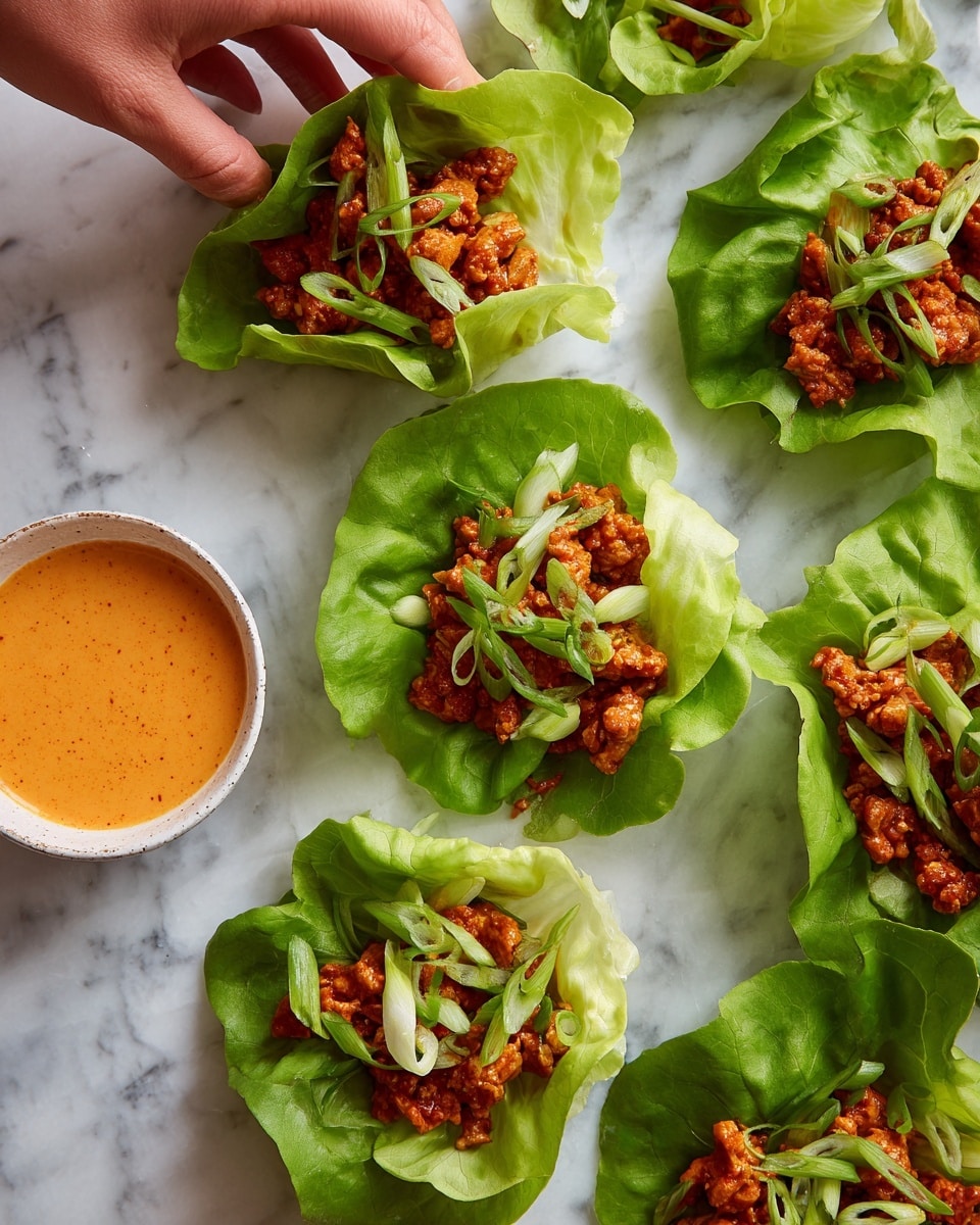 The image shows several green lettuce cups arranged on a white marbled surface. Each cup holds a mixture of small, cooked pieces of meat with a reddish-brown color, topped with thin slices of green onion. The lettuce leaves are fresh and bright light green, cradling the filling with soft, ruffled edges. To the side, there is a small white bowl filled with a thick, creamy orange sauce. A woman's hand is reaching in from the top left corner, holding one of the lettuce cups. The overall look is fresh and colorful with a mix of soft and textured layers. photo taken with an iphone --ar 4:5 --v 7