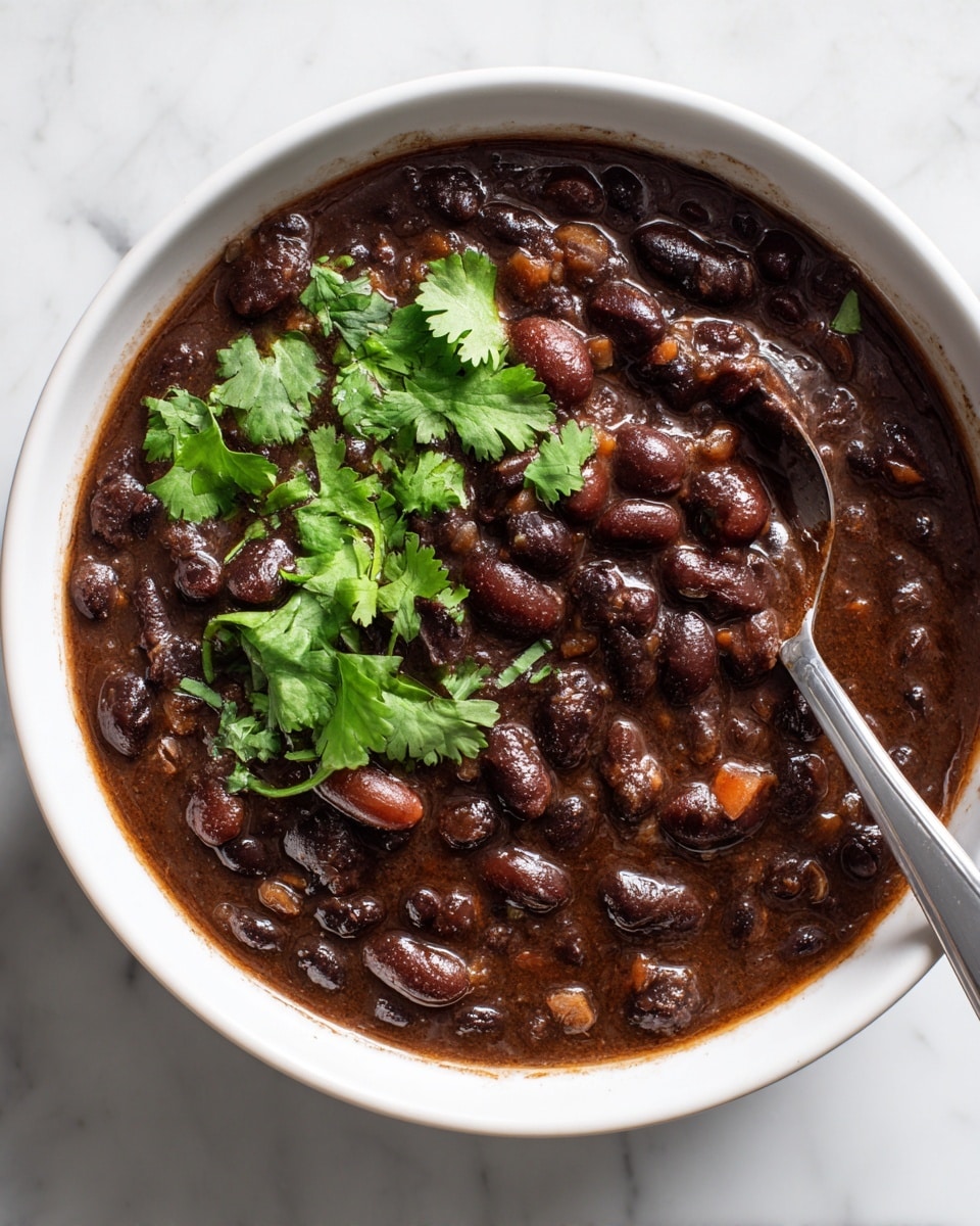 A white bowl filled with thick dark brown beans stew that has a shiny surface with visible whole beans and small pieces inside. On top, there are several fresh green cilantro leaves placed as garnish. A silver spoon rests inside the bowl submerged in the stew, and the bowl is set on a white marbled surface. The edges of the bowl show some sauce stains, adding a natural and warm feel to the dish photo taken with an iphone --ar 4:5 --v 7