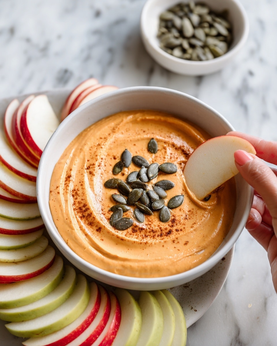 A white round bowl filled with smooth, thick orange dip topped with dark green pumpkin seeds and a light sprinkle of brown spice in the center; around the bowl are layers of apple slices arranged in a circle—green slices, red slices, and pale yellow slices with smooth textures and fresh look; a woman's hand is shown dipping a red apple slice into the orange dip; in the background, there is a small white bowl filled with pumpkin seeds on a white marbled surface. photo taken with an iphone --ar 4:5 --v 7