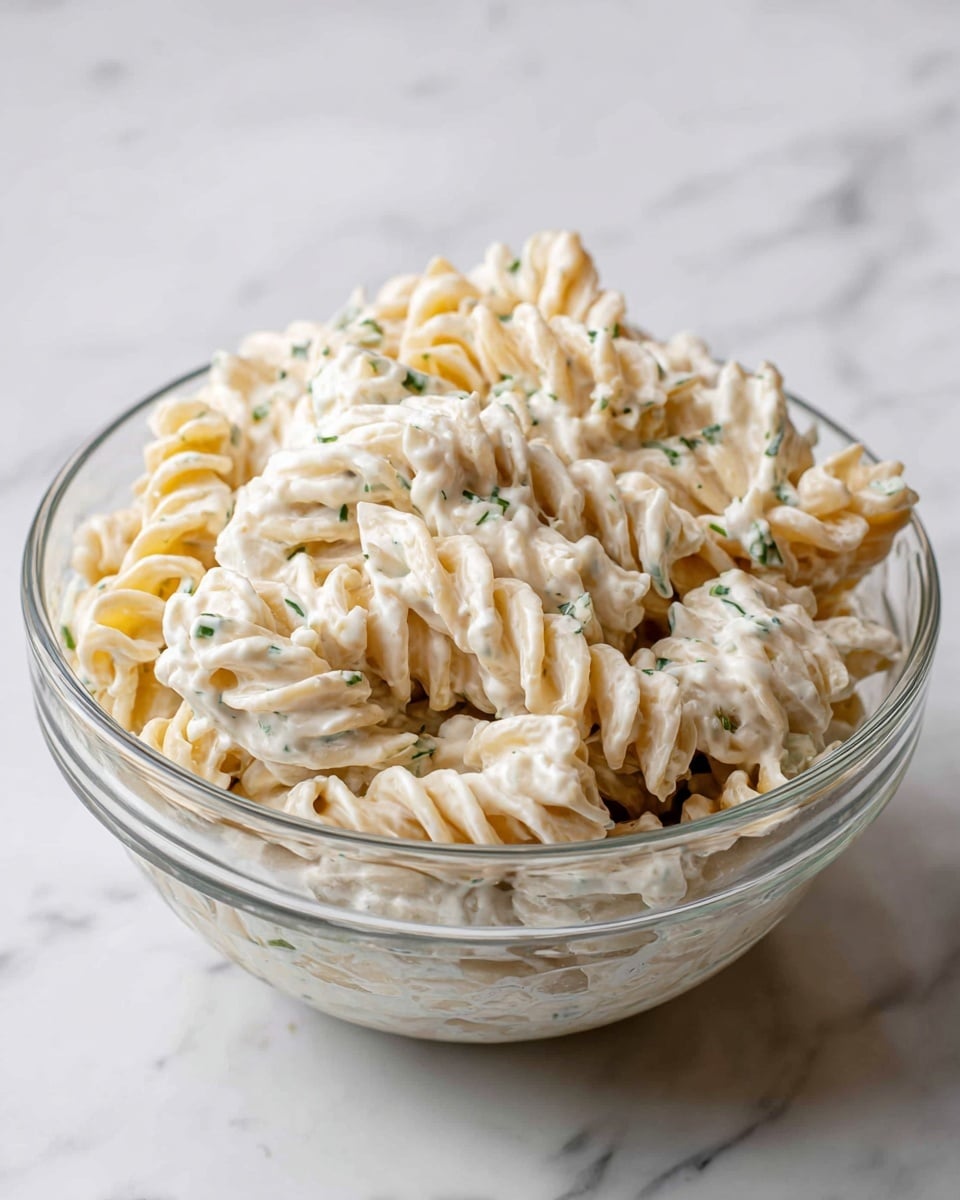 A clear glass bowl filled with three layers of creamy white pasta salad is placed on a white marbled surface. The pasta consists of light yellow twisted noodles coated evenly in a smooth, thick white dressing with small bits of green herbs scattered throughout. The bowl shows some streaks of dressing on the inner sides, and the overall texture looks creamy and mixed fully with the noodles. Photo taken with an iphone --ar 4:5 --v 7