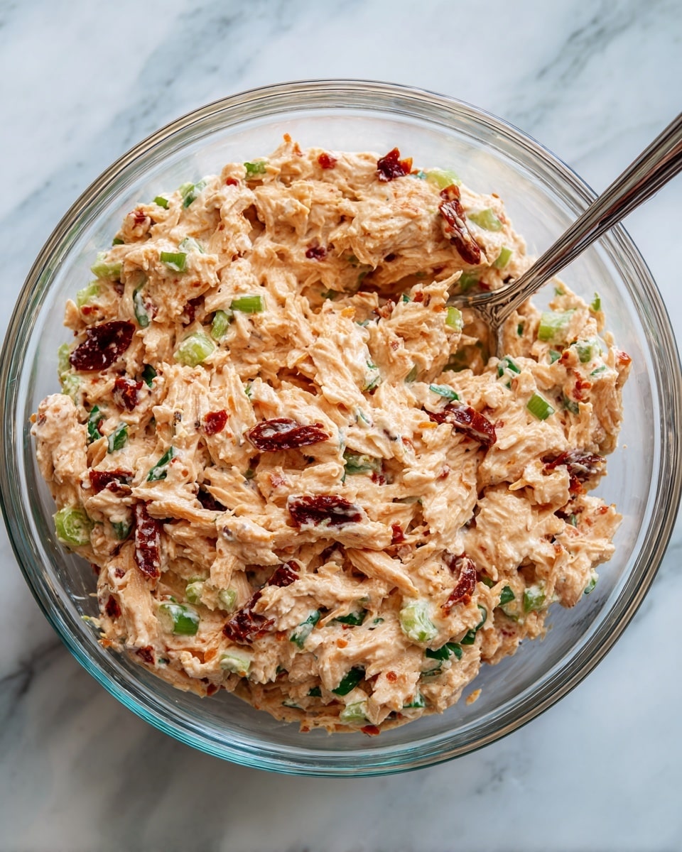 A clear glass bowl filled with a mixed salad that has a creamy, light orange base with visible bits of shredded white meat, bright green celery pieces, dark green leafy herbs, small chunks of dark red sun-dried tomatoes, and finely chopped red onions spread evenly throughout. The mixture looks thick and chunky with a textured appearance, and a metal spoon is partially submerged on the right side. The bowl rests on a white marbled surface. photo taken with an iphone --ar 4:5 --v 7