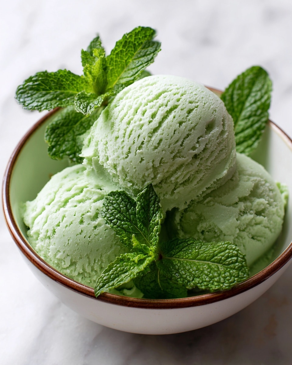 The image shows three scoops of pale green ice cream with a creamy, smooth but slightly textured surface. They sit closely together inside a white bowl with a brown outer rim. Bright green fresh mint leaves with visible veins lay on top and slightly between the scoops. The bowl is placed on a white marbled surface. The scene looks fresh and calming, with cool and natural colors. photo taken with an iphone --ar 4:5 --v 7