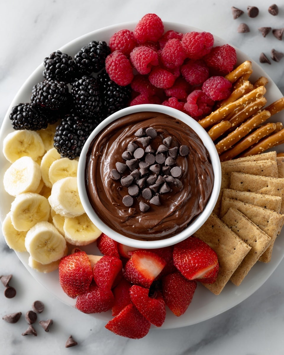 The image shows a white round plate with a white small bowl in the center filled with thick, dark brown chocolate dip topped with scattered dark chocolate chips. Surrounding the bowl are fresh blackberries on the top left, bright red raspberries on the top right, sliced red strawberries on the bottom right, light yellow banana chips on the bottom left, golden brown graham crackers on the left, and golden brown pretzels on the right. The plate is placed on a white marbled surface with a few chocolate chips scattered in the background. Photo taken with an iphone --ar 4:5 --v 7