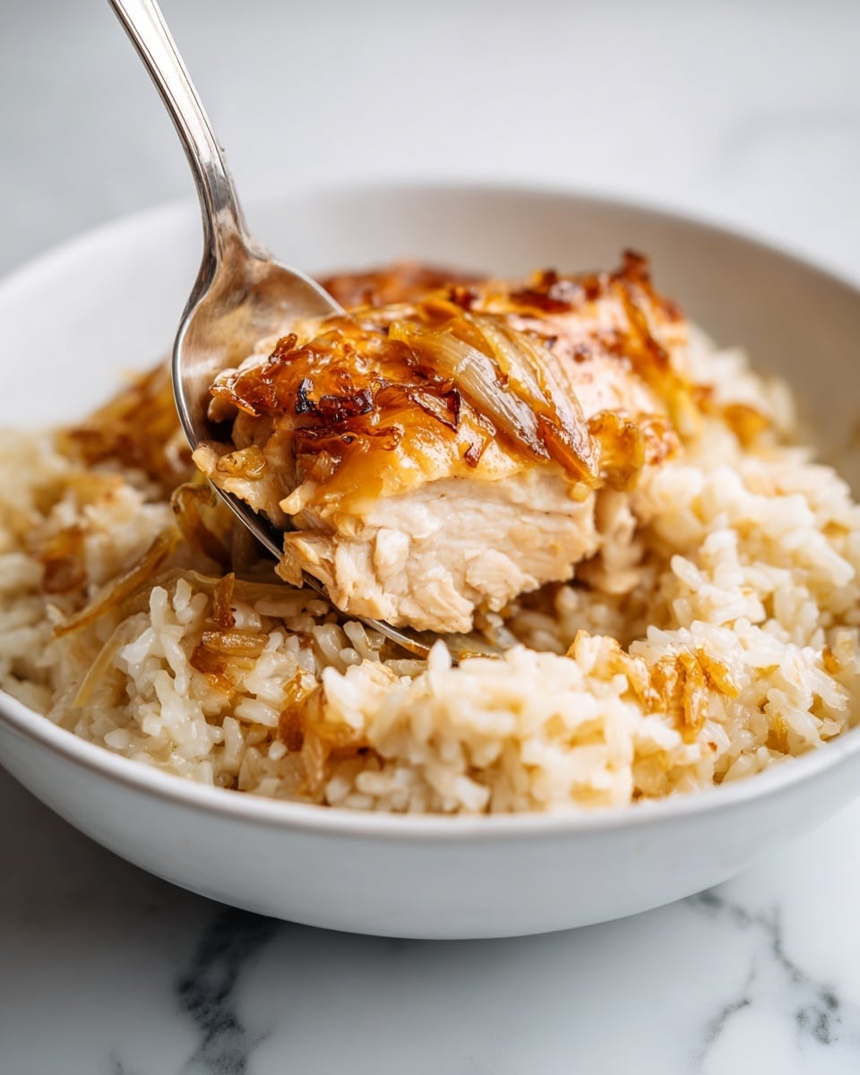 A close-up of a dish in a white bowl showing two main layers: on the top, a piece of cooked chicken with a golden-brown, slightly shiny and textured surface with bits of caramelized onions, and below it, a creamy rice layer that is beige with a soft, moist texture, speckled lightly with small bits of onion or seasoning. A metal spoon is scooping the rice near the chicken, all set on a white marbled surface. photo taken with an iphone --ar 4:5 --v 7