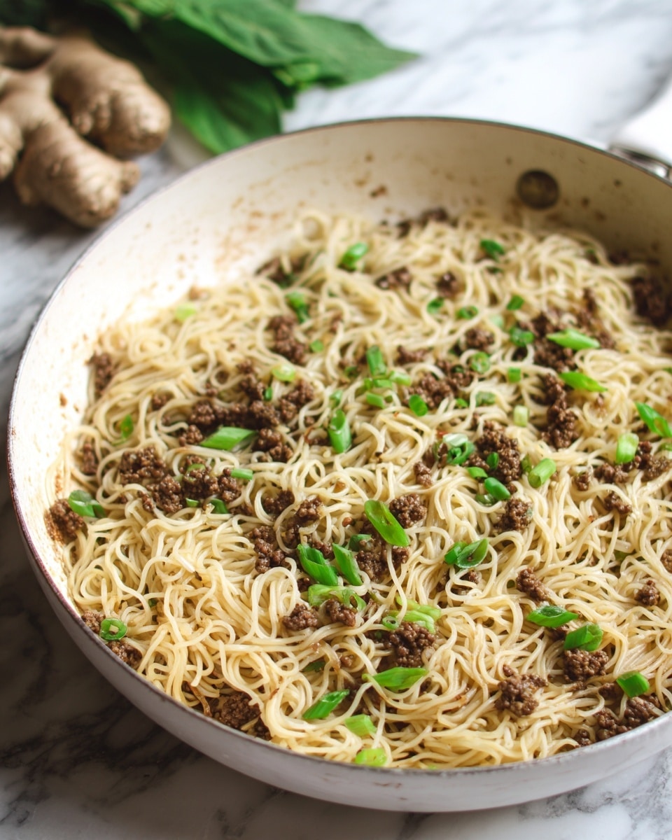 The dish is a skillet filled with a mix of noodles and browned minced meat, scattered with small green onion pieces on top. The noodles are light beige and look soft, tangled throughout the pan. The meat is finely ground and dark brown, mixed evenly with the noodles. The skillet is white with slight browning marks on the inside edges. The background is a white marbled surface, with some ginger root and green leaves blurred in the distance. Photo taken with an iphone --ar 4:5 --v 7