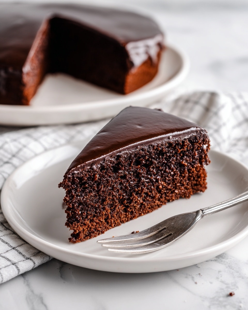 A single slice of chocolate cake with two layers is placed on a white plate. The bottom layer is dark brown and moist with a slightly crumbly texture, while the top layer is a smooth, glossy chocolate glaze that covers the entire surface of the cake slice. The cake is angled with its pointed tip facing the front. To the right side of the plate, there is a silver fork lying flat. In the background, a larger white plate holds the rest of the cake with one slice missing. The surface underneath the plates is a white marbled texture, and a white and gray checkered cloth is partly visible under the plate with the cake slice. Photo taken with an iphone --ar 4:5 --v 7