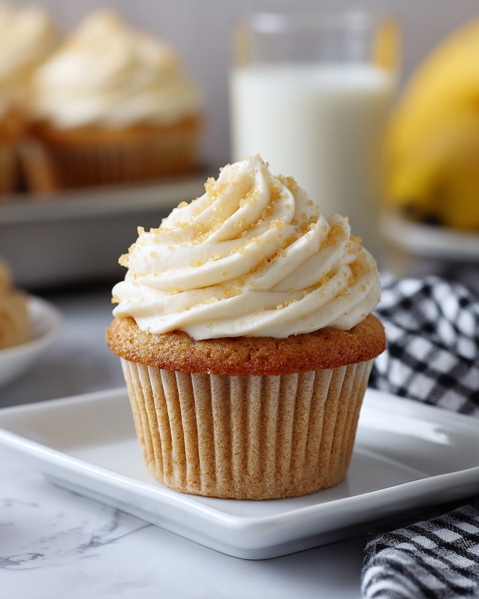 A single cupcake sits in the center of a white square plate on a white marbled surface. The cupcake has one visible layer of light brown cake with a textured, slightly rough surface, wrapped in a ridged paper liner. On top, there is a thick, swirled layer of creamy white frosting sprinkled with small, light golden sugar crystals. The background shows a blurred glass of milk and a banana, along with a black-and-white checkered cloth and a tray holding more cupcakes. Photo taken with an iphone --ar 4:5 --v 7