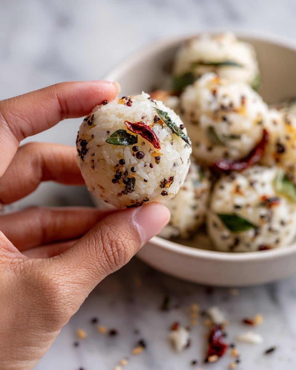 A close-up view shows a woman's hand shaping a small, oval white rice ball with tiny black mustard seeds, reddish pieces, and green herbs embedded in it. The rice ball has a soft, grainy texture. Behind, there is a white bowl filled with more of this white rice mixture, scattered with dark mustard seeds, red chili pieces, and green leaves. The scene rests on a white marbled surface. photo taken with an iphone --ar 4:5 --v 7