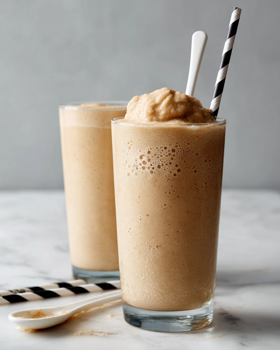 Two clear glasses filled with thick, light brown milkshake with a frothy, bubbly texture on top. The glass in the front has a white plastic spoon lifting a dense scoop of the milkshake above the glass rim, and a black-and-white striped paper straw leaning inside the glass. The second glass behind it also shows a bubbly surface with a similar striped straw. Four more black-and-white striped straws lie scattered on a white marbled surface, which also holds a white ceramic spoon with some milkshake residue. Photo taken with an iphone --ar 4:5 --v 7