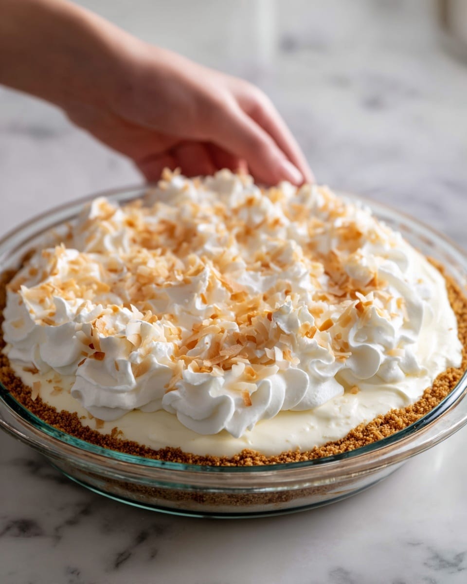 The image shows a clear glass pie dish filled with a thick, evenly pressed light brown cookie crust layer at the bottom. Above the crust, there is a creamy white pudding layer, smooth and glossy, topped with a fluffy layer of bright white whipped topping that looks soft and airy. Scattered lightly on top of the whipped layer are small pieces of toasted coconut, golden brown in color with a slightly crispy texture. Each layer is distinct and neatly arranged, set on a white marbled surface. A woman's hand is poised near the pie, as if about to serve or finish the dessert. photo taken with an iphone --ar 4:5 --v 7