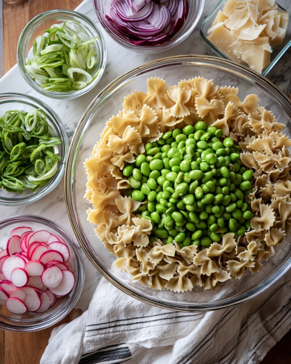 A large clear glass bowl sits in the center filled with three layers: bottom layer of beige bow-tie pasta, a middle layer of bright green edamame beans, and a top layer thickly covered with fresh, glossy green peas. Around the bowl, multiple small clear and white bowls hold vibrant green sliced scallions, light pink thinly sliced radishes, thin light purple onion rings, and beige artichoke pieces. A container of shaved Parmesan sits in the top right corner. The scene is set on a wooden table with a white marbled texture and a white cloth with dark stripes partially under the main bowl. Photo taken with an iphone --ar 4:5 --v 7