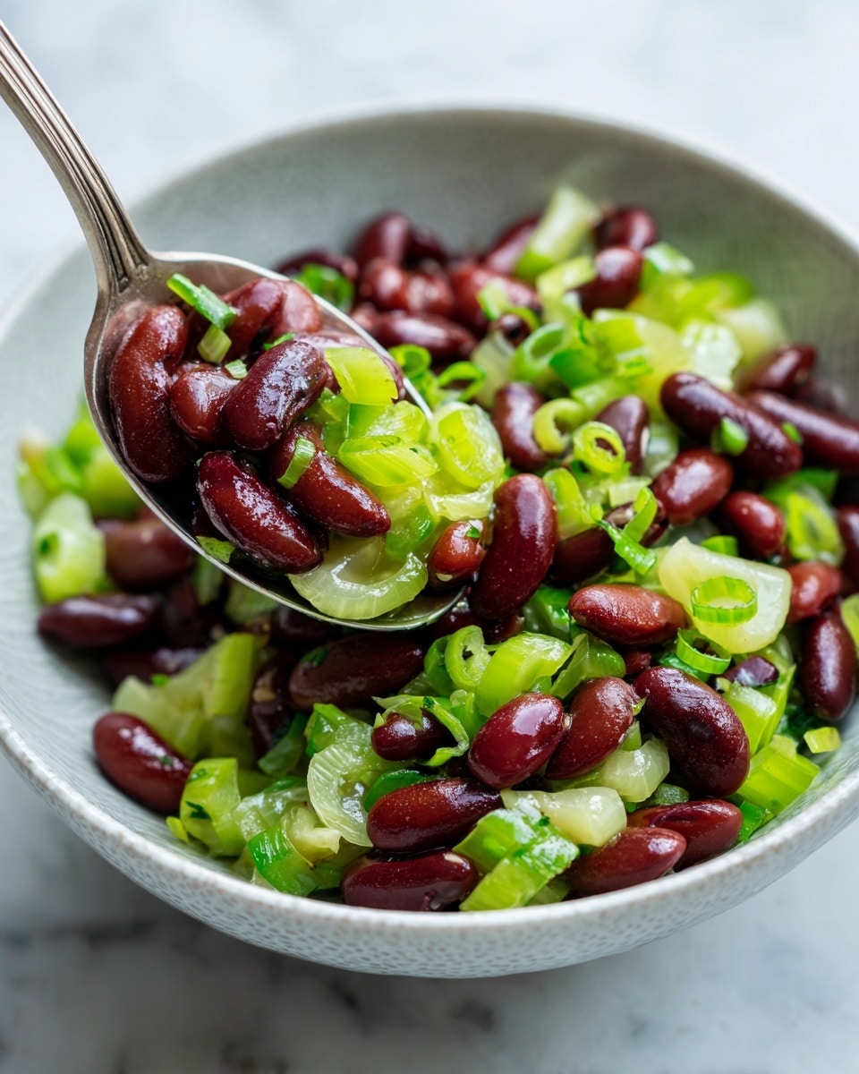 The image shows a close-up of a bowl filled with a salad made of dark red kidney beans and light green celery slices. The beans are shiny and smooth, scattered evenly throughout the salad, adding a rich red color. The celery slices add a fresh light green color with a slightly crunchy texture, and you can see small rings of green onion mixed in, adding a touch of brightness. The salad looks creamy, covered lightly with a dressing. The bowl is white with a subtle pattern, placed on a white marbled surface, and a spoon is scooping part of the salad upwards. photo taken with an iphone --ar 4:5 --v 7