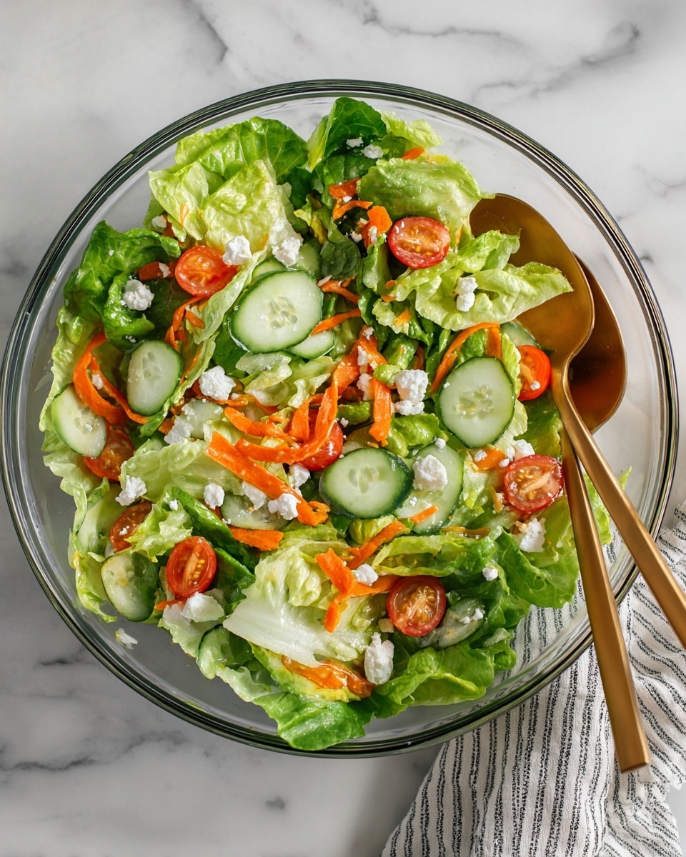 A clear glass bowl holds a fresh mixed salad with large green lettuce leaves forming the base layer, topped with round slices of light green cucumber, small bright red cherry tomato halves, and thin, curly orange carrot strips scattered throughout. White soft cheese crumbles are sprinkled evenly over the salad, adding texture and contrast. Two gold-colored serving spoons rest on the side inside the bowl, inviting to serve. The bowl sits on a white marbled surface with a glimpse of a striped cloth napkin on the right edge. Photo taken with an iphone --ar 4:5 --v 7