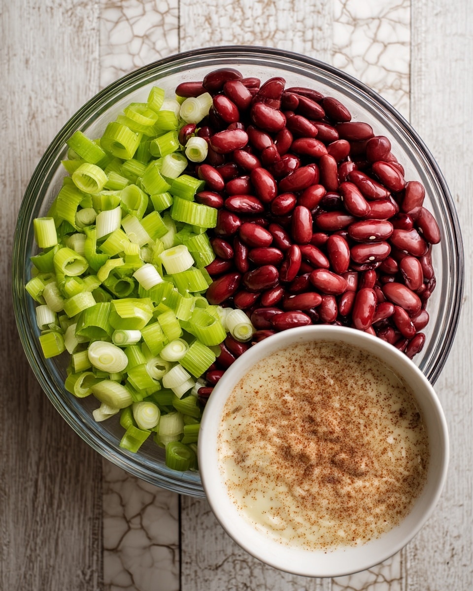 A clear glass mixing bowl filled with three main ingredients: bright red kidney beans covering the right half, fresh light green celery sliced into thin crescent shapes on the top left, and chopped green onions with white and green rings scattered along the bottom left. Below this bowl, there is a white bowl containing a creamy mixture topped with a sprinkle of light brown spice or seasoning. The setting is on a wooden surface with a white marbled texture background. Photo taken with an iphone --ar 4:5 --v 7