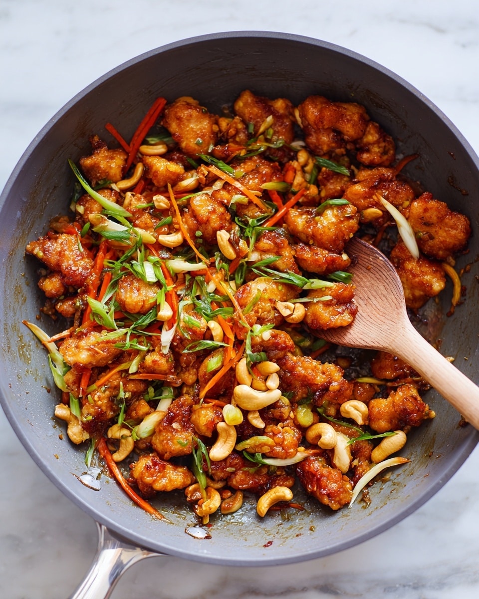 A grey cooking pan filled with a mix of small crispy brown chicken pieces, thin orange carrot strips, light brown cashew nuts, and chopped green onions scattered throughout, with a wooden spoon resting inside. The chicken pieces have a crunchy texture and are evenly coated in sauce, while the carrots are fresh and slightly shiny. The green onions add a fresh contrast, and the cashews provide a smooth, light-colored detail. The pan sits on a white marbled surface. photo taken with an iphone --ar 4:5 --v 7