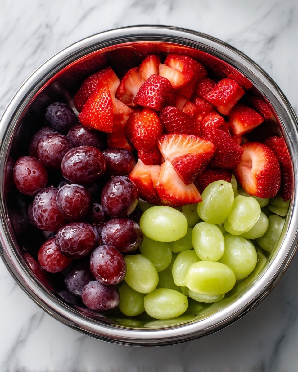A shiny silver bowl contains three sections of fresh fruit neatly separated. On the left, there are deep purple grapes, some whole and some cut to show their juicy inside. In the middle, bright red strawberries are sliced into small pieces, showing their white inner flesh and seeds on the outside. On the right, light green grapes are also halved, revealing their shiny, smooth inside. The background shows a white marbled texture. photo taken with an iphone --ar 4:5 --v 7