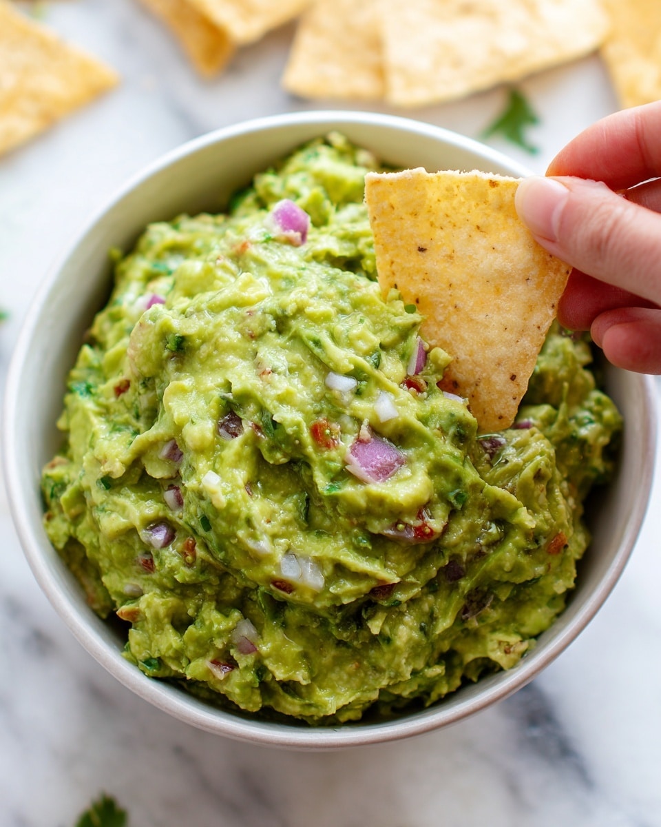 A close-up of a white bowl filled with chunky green guacamole that has visible pieces of purple onion and small bits of red tomato mixed in. A woman's hand is holding an off-white, triangular tortilla chip dipped into the guacamole, lifting it slightly above the bowl. The background shows some blurred chips and green leaves on a white marbled surface. The colors are bright and fresh, highlighting the creamy texture of the guacamole with small, uneven chunks throughout. photo taken with an iphone --ar 4:5 --v 7