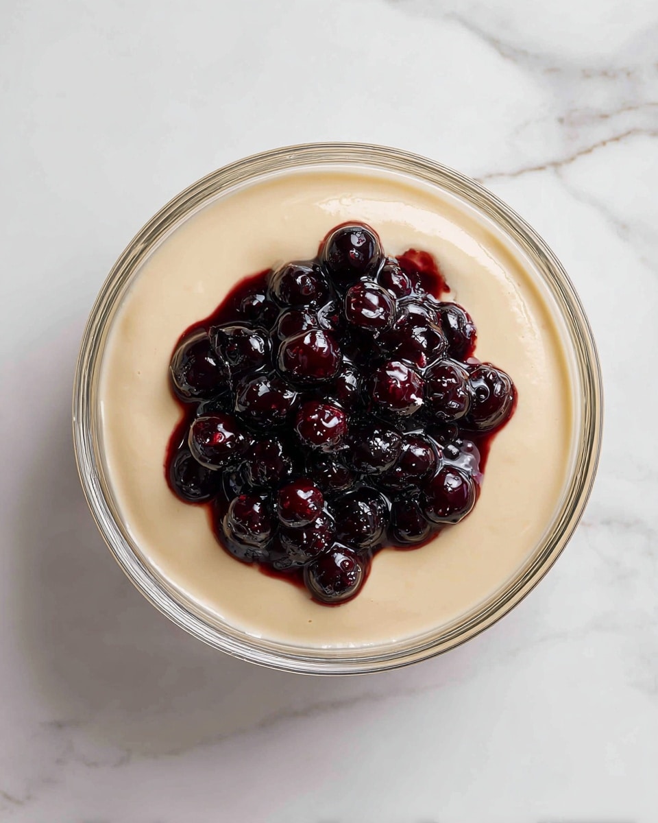 A clear glass bowl is filled with a smooth, light beige layer that looks creamy and thick, covering the entire base. In the center on top, there is a pile of glossy, dark purple mixed berries in syrup, with individual berries visible and some syrup shining under the light, creating a rich contrast with the beige layer around it. The bowl is set against a white marbled surface, enhancing the colors of the dish. photo taken with an iphone --ar 4:5 --v 7