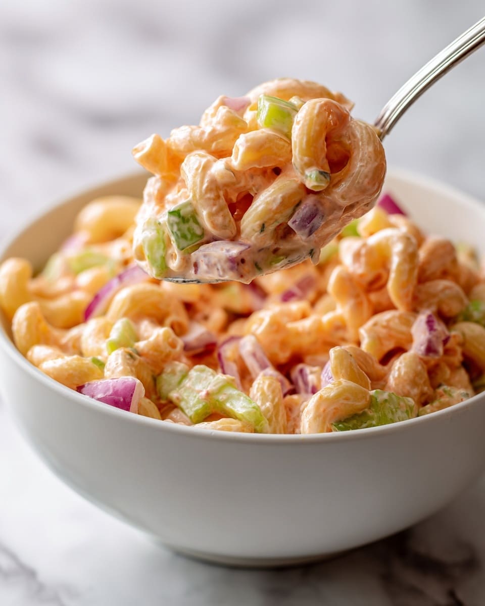 This image shows a close-up of a white bowl filled with a creamy pasta salad. The salad has three main layers: small curved macaroni pasta coated in a shiny pinkish-orange dressing, small chunks of green celery scattered throughout, and thin slices of purple-red onion mixed in. A silver spoon holding a portion of the salad is lifted above the bowl, showing the creamy texture and mixed ingredients clearly. The background is softly blurred with a white marbled texture. Photo taken with an iphone --ar 4:5 --v 7