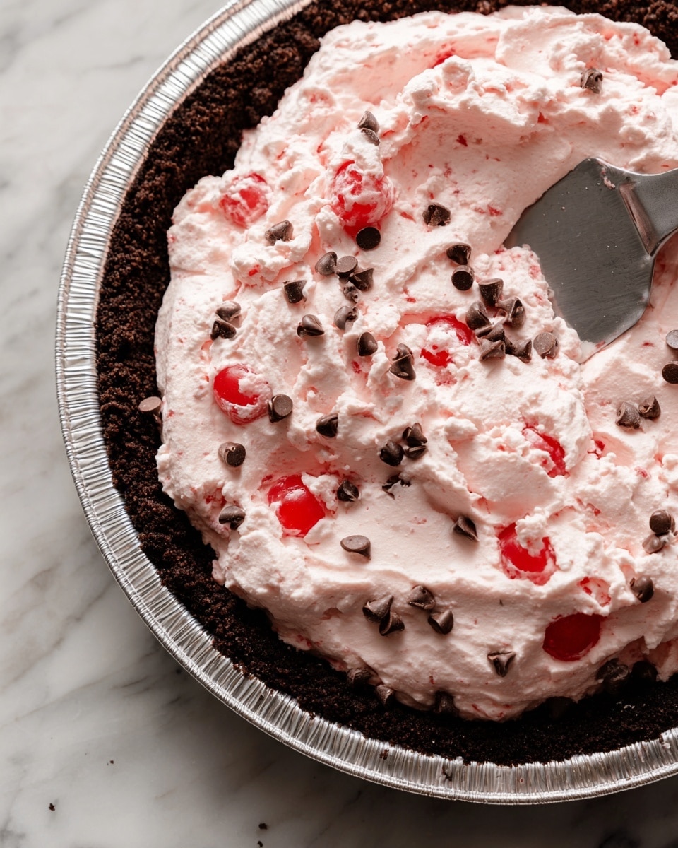 A close-up view shows a pie with two main layers on a white marbled surface. The bottom layer is a dark brown, almost black, crumbly crust inside a silver foil pie pan. The top layer is a thick, pale pink cream mixed with small dark chocolate chips and bright red cherry pieces, spread evenly over the crust. A metal spatula is partially visible, smoothing the pink filling across the surface. In the top left corner, a smaller inset shows the same pie from above, with the pink filling still fluffy and being spread inside the crust. Photo taken with an iphone --ar 4:5 --v 7