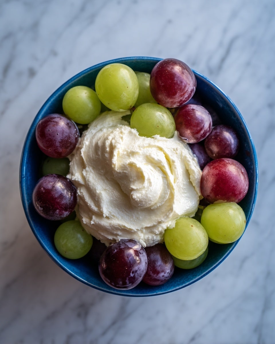 A close-up image of a blue bowl filled with two layers: the bottom layer is made up of whole green and purple grapes, and the top layer is a thick, creamy white spread placed roughly in the center of the grapes. The bowl is on a white marbled surface. photo taken with an iphone --ar 4:5 --v 7